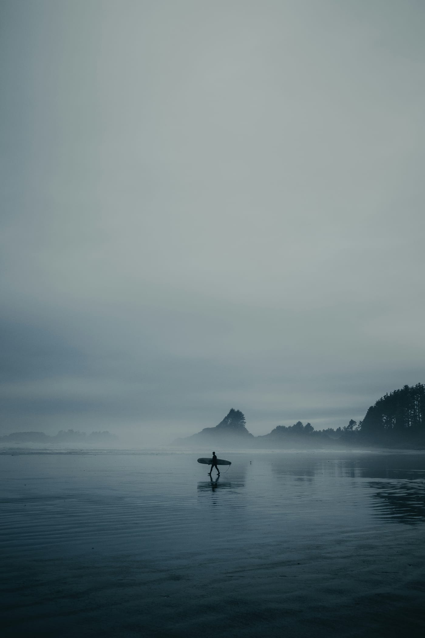 Surfer walking along a misty Tofino beach with surfboard in winter